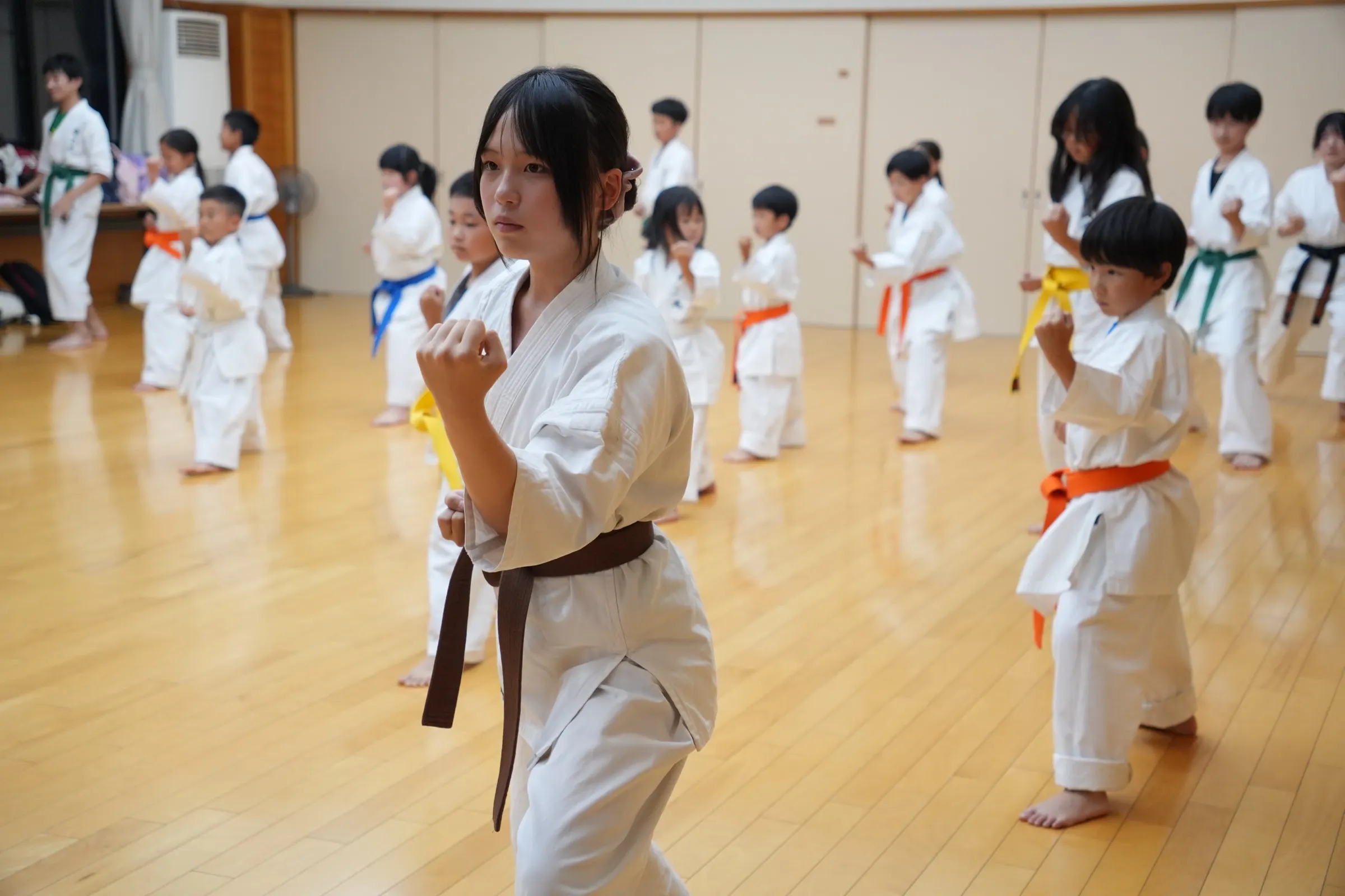 指導者と子どもたちが背景の生徒たちと一緒に稽古に取り組む様子
